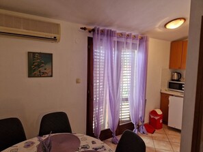 A room with a dining table, chairs, a kitchen area with a microwave, and a window with purple curtains.
