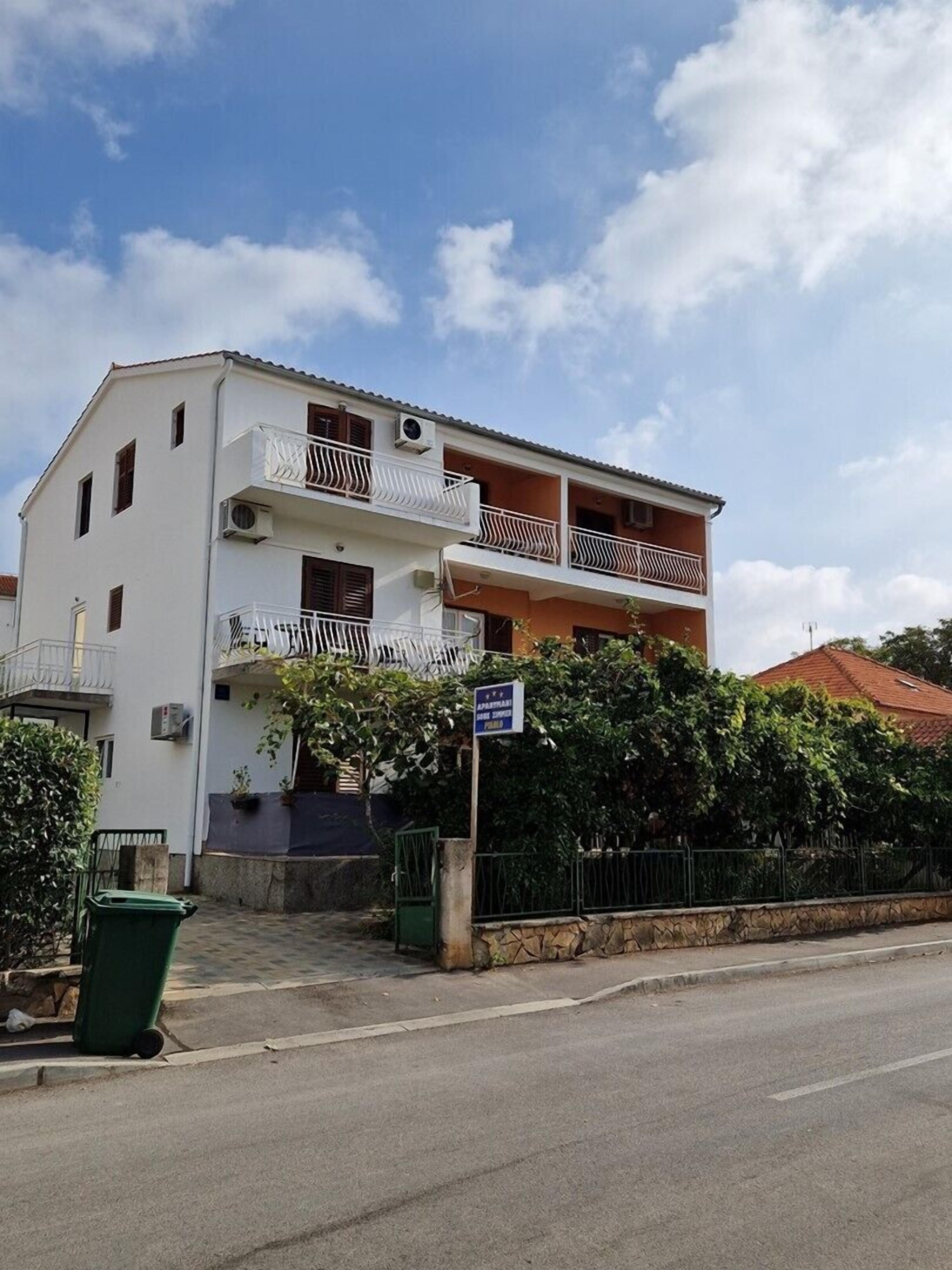 Un edificio de varios pisos con balcones, un bote de basura verde y un letrero que indica un establecimiento público.