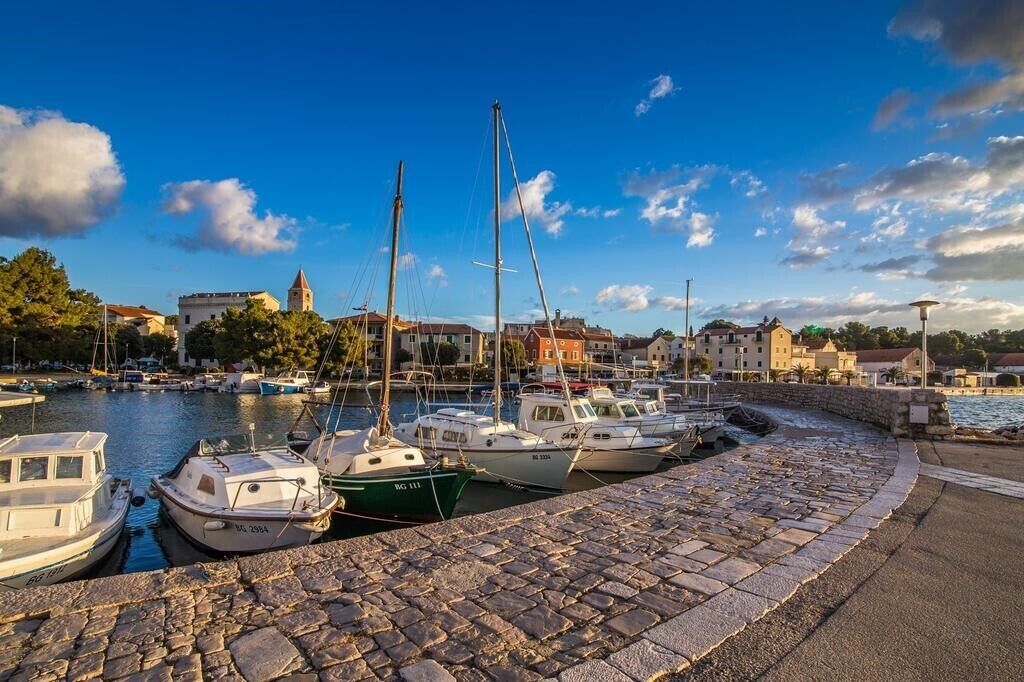 En småbåtshamn med förtöjda båtar, en stenbelagd gångväg och byggnader i bakgrunden.