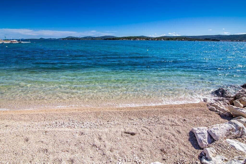 Une plage dégagée, avec des galets et des rochers, une eau calme et des collines qui se dessinent à l’horizon.