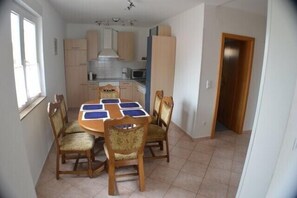 A dining area with a round table and six chairs, a kitchen in the background, and a wooden door.