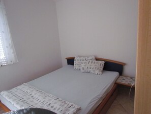 Une petite chambre simple, avec un lit simple, une table de chevet et une fenêtre ornée d’un rideau léger.