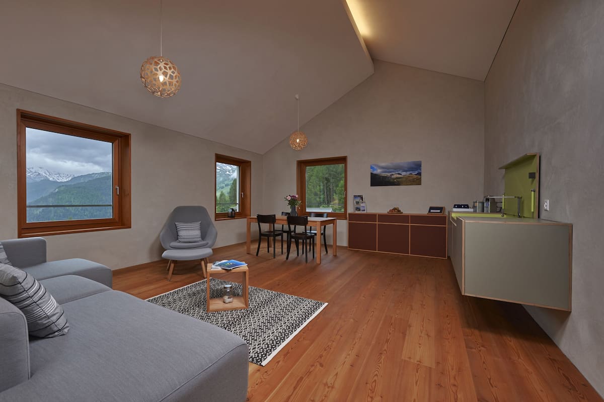 Ein modernes Wohnzimmer mit einer Couch, einem Esstisch und Stühlen. Große Fenster bieten Blick auf die Berge.