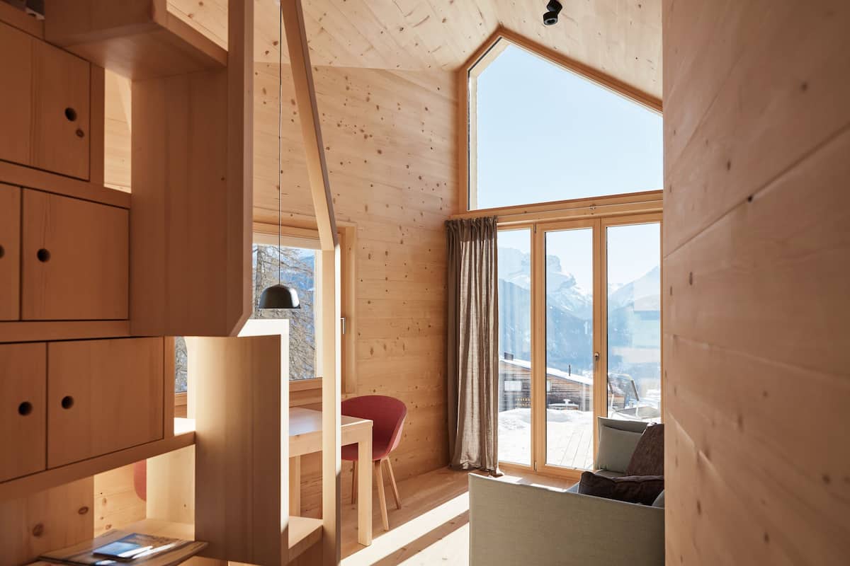 Innenansicht einer Holzhütte mit großem Fenster, Holzwänden, einem Essbereich und Blick auf einen verschneiten Berg.