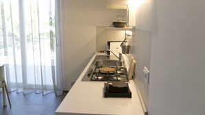 A compact kitchen with a gas stove, a sink, and a small countertop. There is a window with sheer curtains, and a chair is visible to the left. - Via del Faro Apartments - BUSSOLA (Torre Canne)