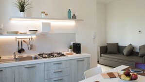 Cocina moderna con estufa a gas, lavamanos y utensilios colgantes. Hay un comedor con una mesa preparada para dos personas, un sofá y un frutero.