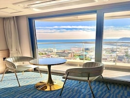 Habitación de hotel con un ventanal que ofrece vistas al mar y al paisaje urbano. Hay una mesa redonda con dos sillas y una alfombra azul.