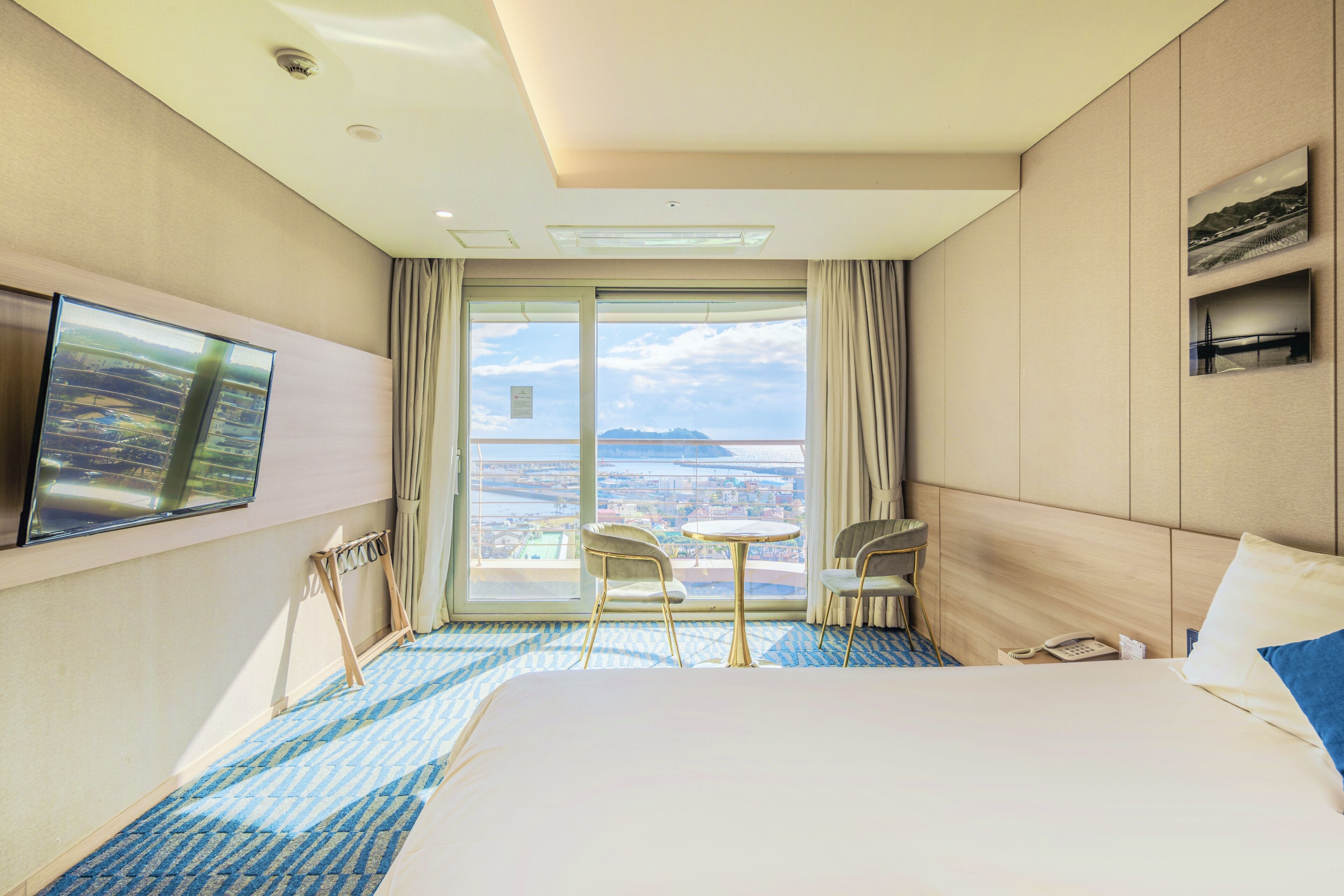A hotel room with a large bed, a small table, two chairs, a TV, and a view of the city.