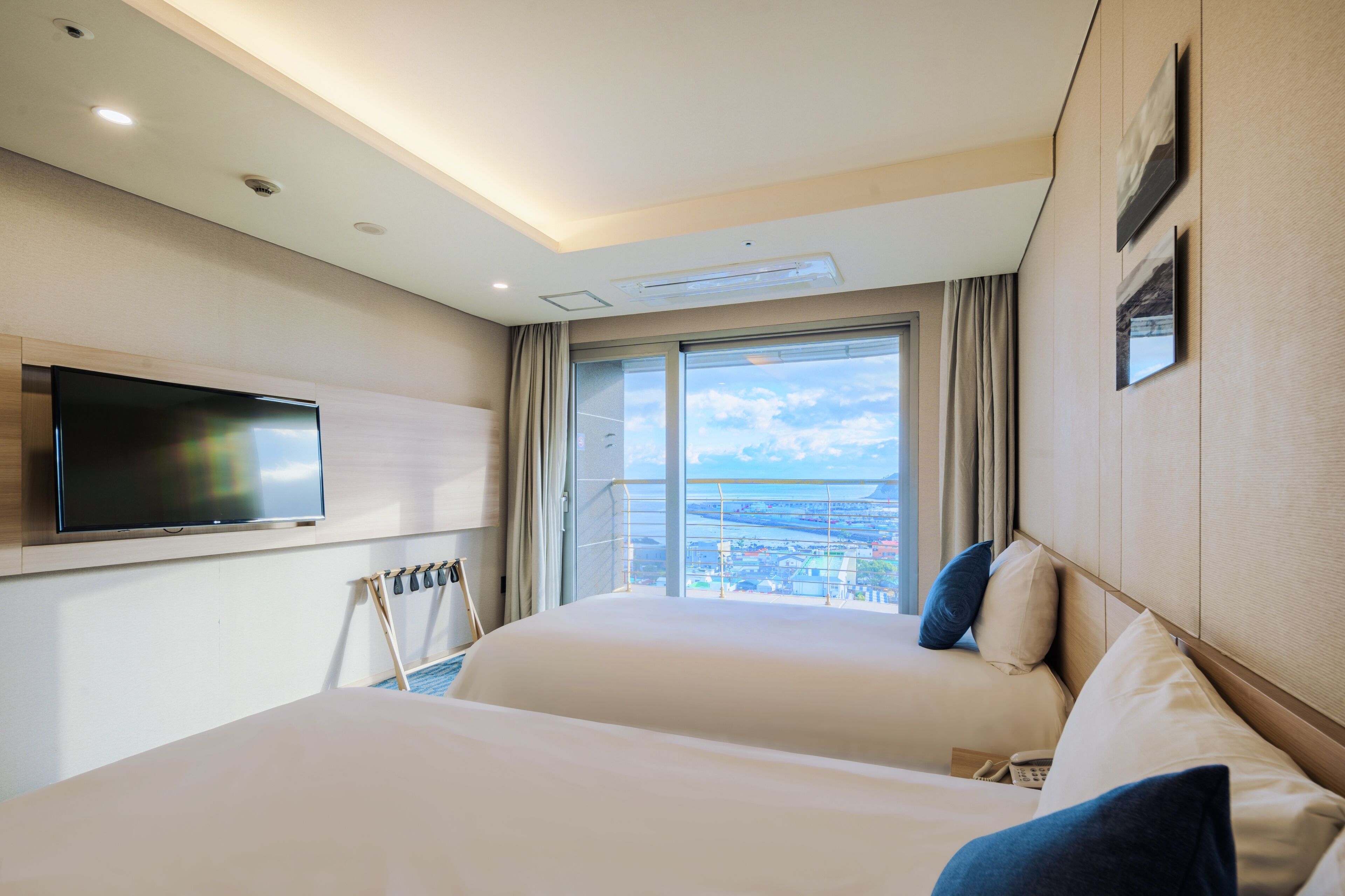 Une chambre d’hôtel avec deux lits, un téléviseur à écran plat fixé au mur, une grande fenêtre donnant sur la ville et un plafond doté d’un éclairage encastré.