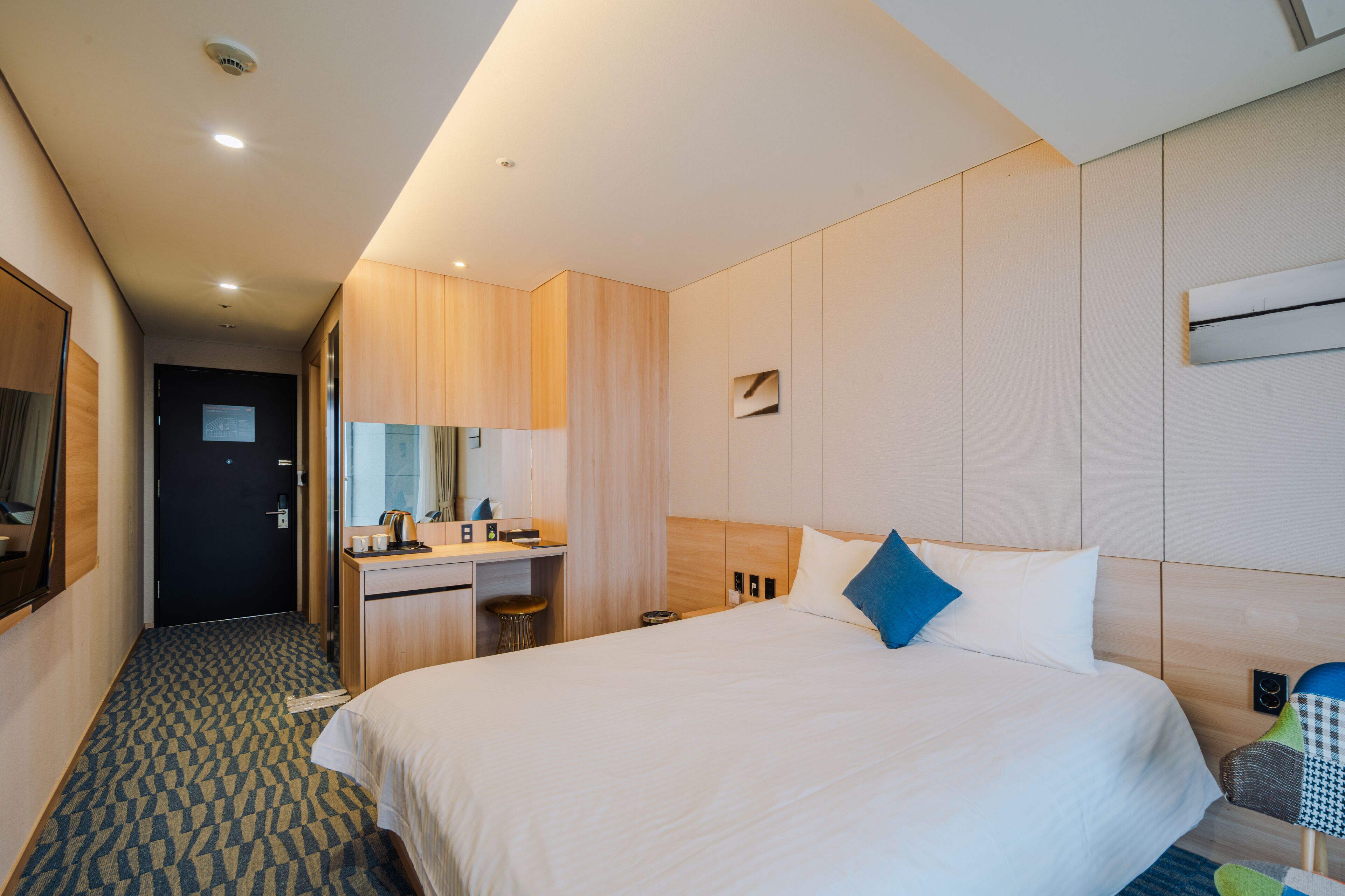 Una habitación de hotel con una cama grande, un escritorio con silla, un espejo y un taburete pequeño.