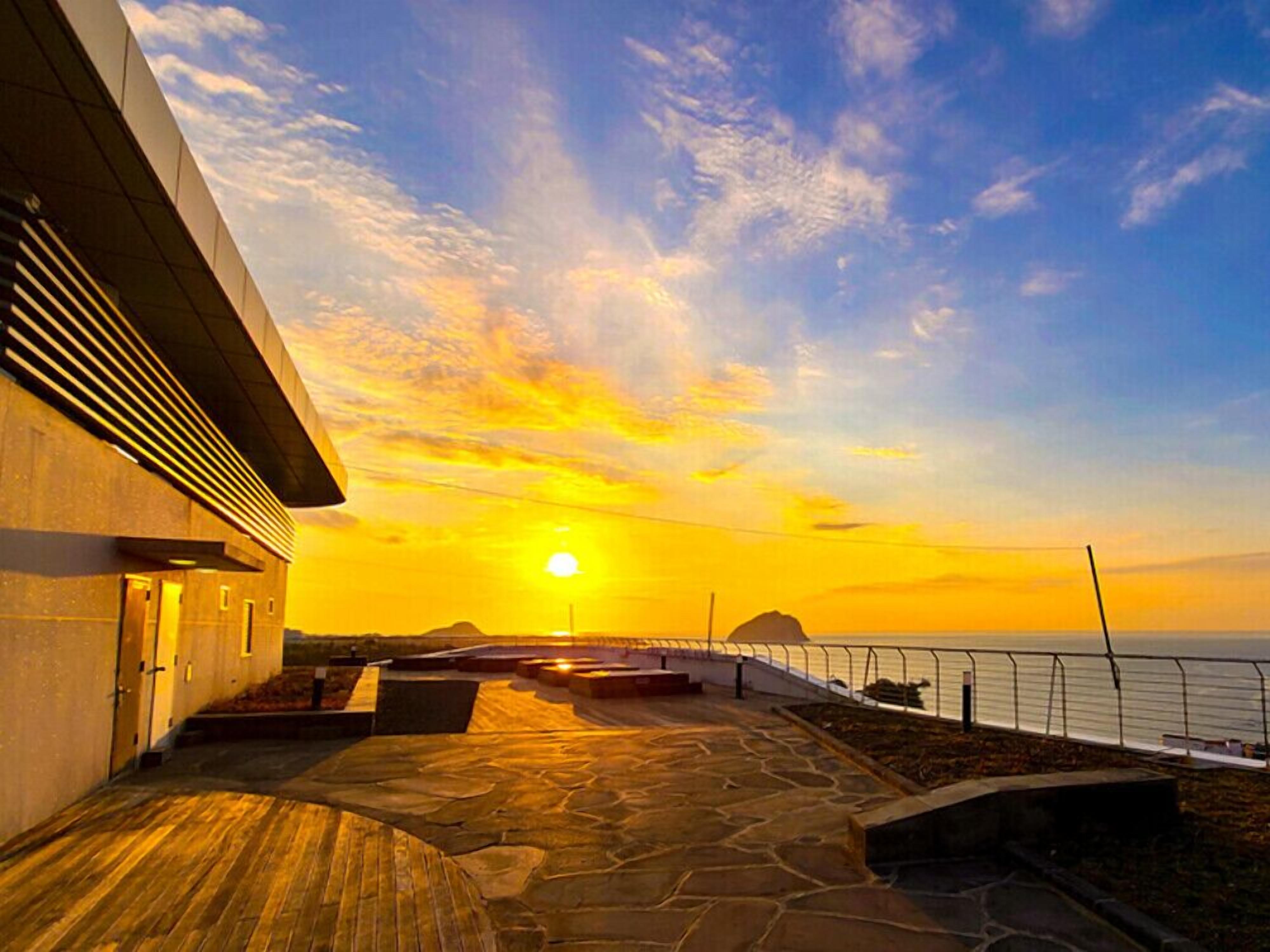 A sunset view from a stone-paved terrace with a modern building on the left and a railing overlooking the sea.