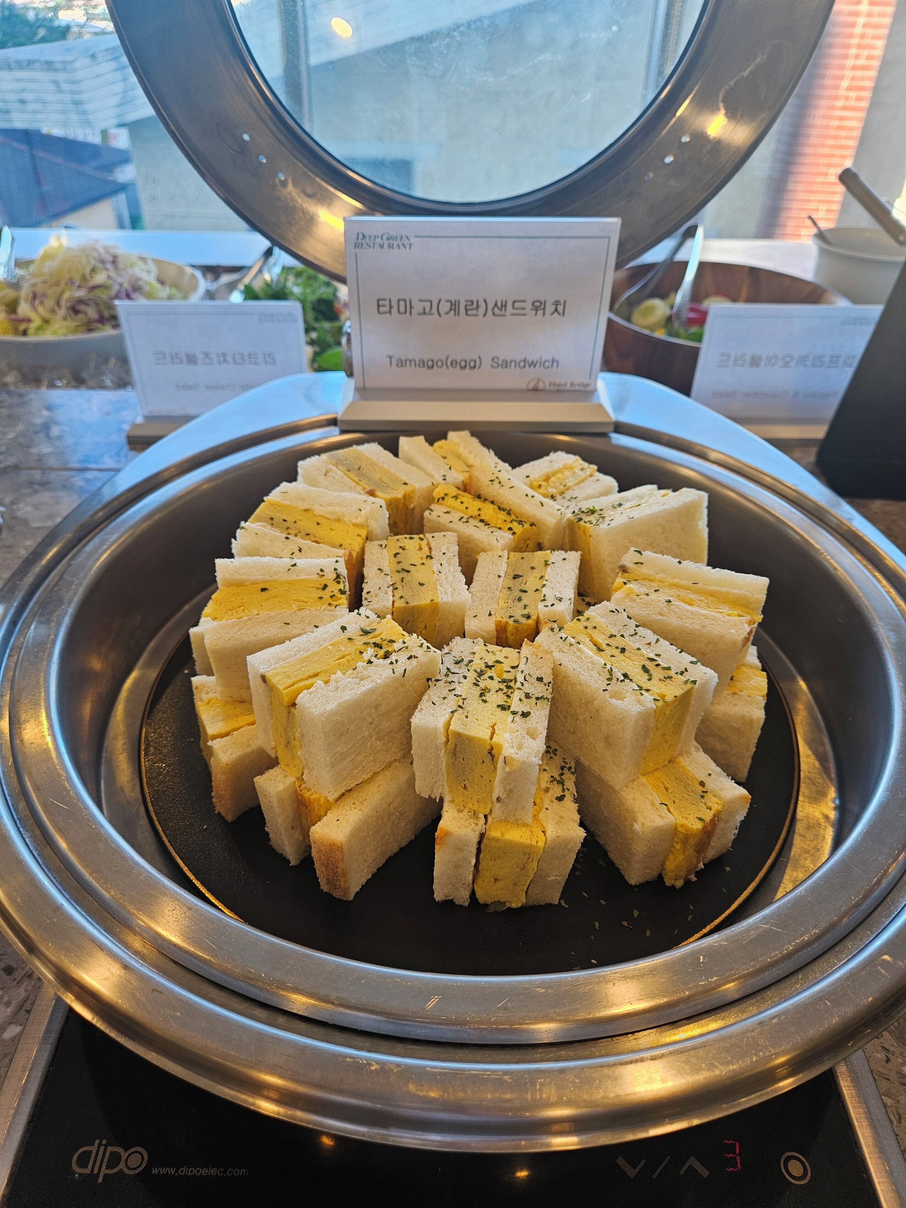 A tray of tamago (egg) sandwiches garnished with herbs, displayed on a metal stand with a sign above.