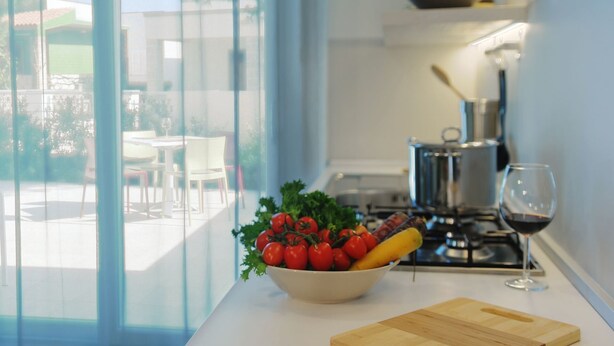 A kitchen with a bowl of fresh tomatoes and a banana on the counter, a cutting board, a wine glass, and a stove. - Via del Faro Apartments - OVEST (Torre Canne)