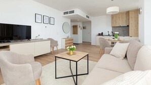 A modern living room with a sofa, armchair, and coffee table. There is a kitchen area with wooden cabinets and a dining table.