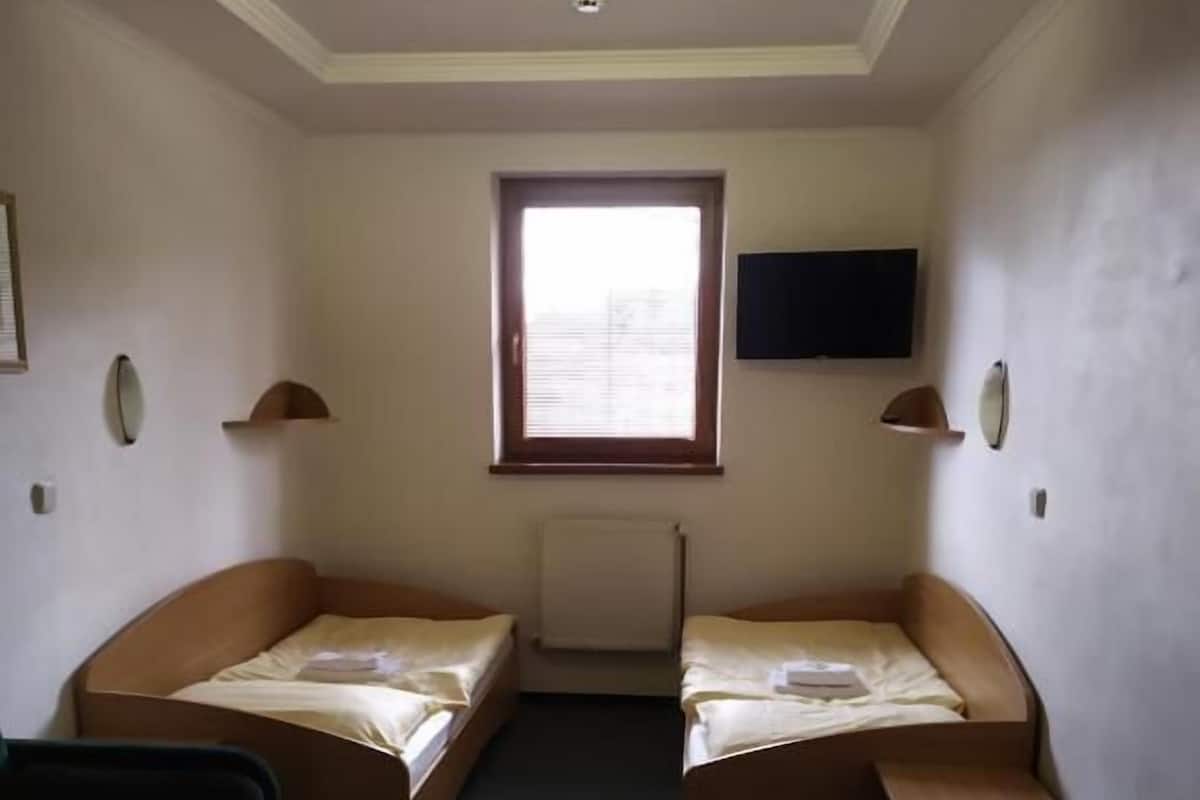 A small, compact room with two single beds, a desk, a chair, and a television mounted on the wall.
