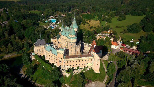 An aerial view of a large castle with multiple towers and a moat, surrounded by lush greenery and a small village. - Penzión DRUŽBA Bojnice (Bojnice)