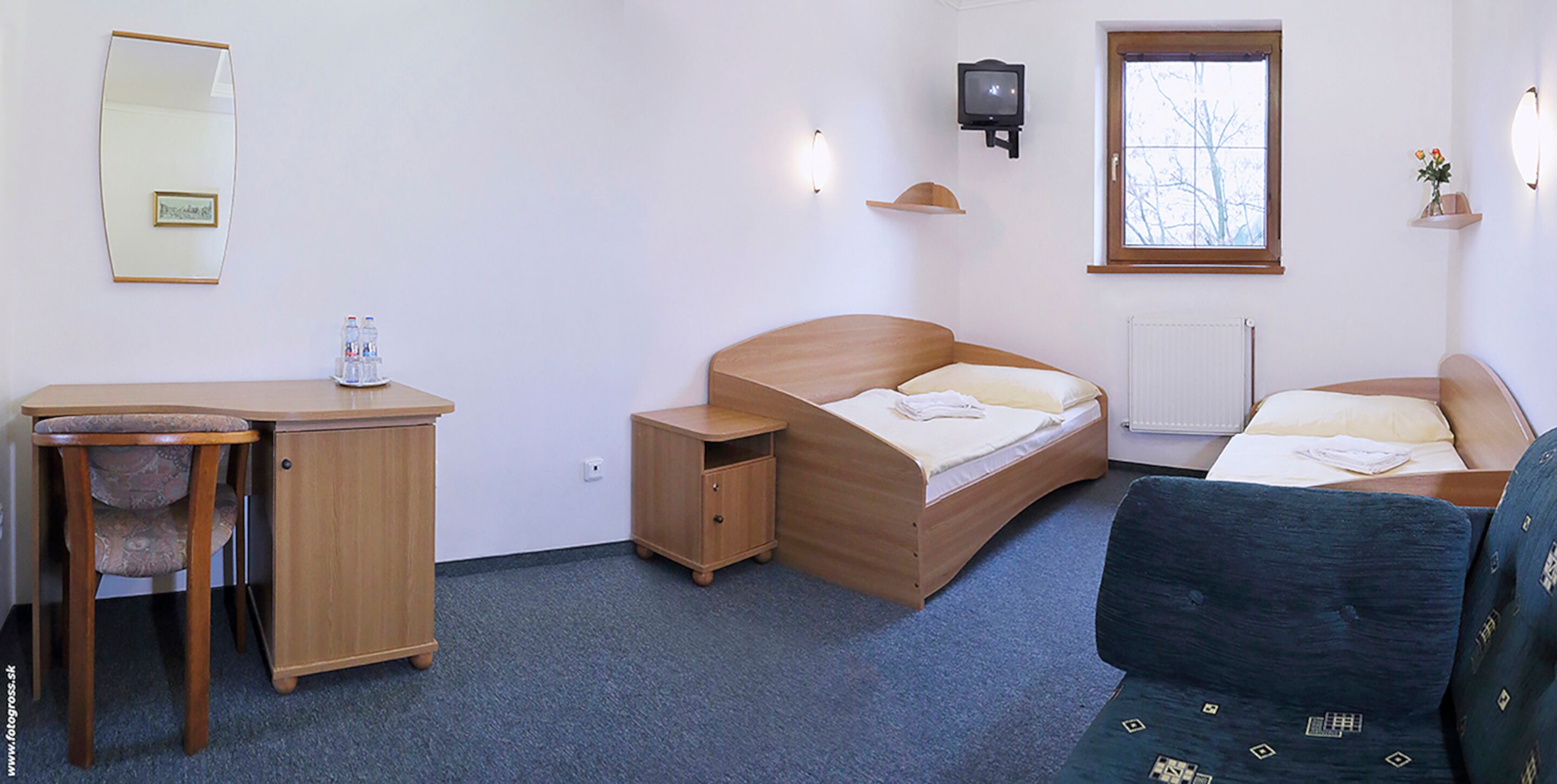 A bedroom with a wooden bed, a desk with a chair, a dresser, a television, and a window with a view.
