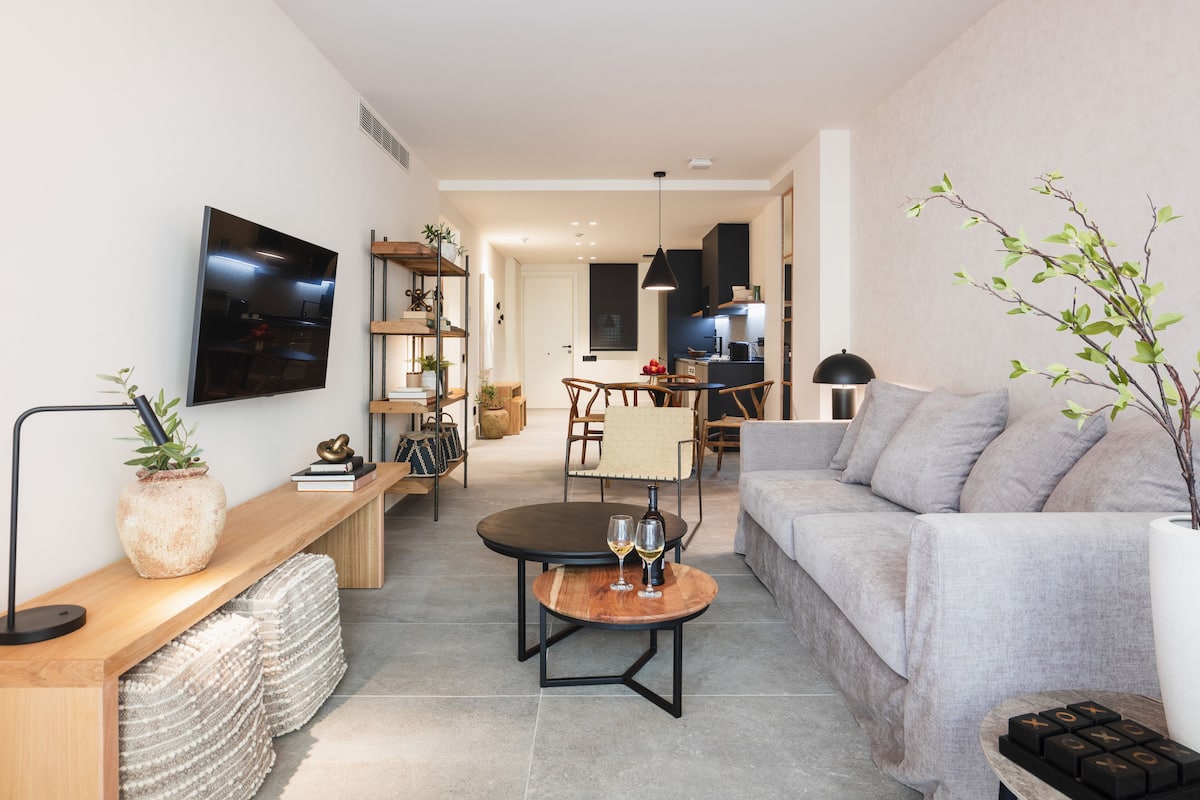 Un salon moderne avec un canapé gris, une table basse sur laquelle reposent deux verres de vin, une table d’appoint en bois agrémentée d’une plante, et un téléviseur fixé au mur.
