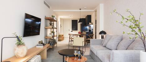 Una sala de estar moderna con un sofá gris, una mesa de centro con dos copas de vino, una mesita de noche de madera con una planta y un televisor montado en la pared.