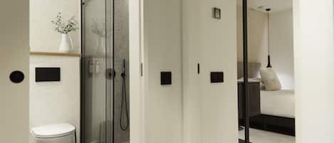 A modern bathroom with a glass shower enclosure, a toilet, and a bedside table with a lamp.