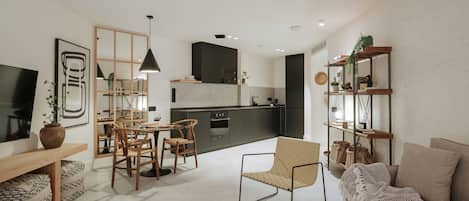 A modern living room with a sofa, a dining area with chairs, a kitchen with dark cabinets, and a TV mounted on the wall.