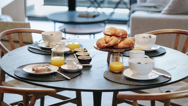 Une table de petit-déjeuner dressée avec des croissants, des bols, des tasses et du jus d’orange, dans une pièce moderne dotée de grandes fenêtres et offrant une vue sur des palmiers.