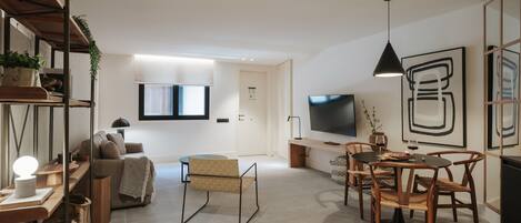 Una sala de estar moderna con un sofá, una mesa de centro, una mesa de comedor con sillas, un televisor montado en la pared y un gran ventanal con persianas.