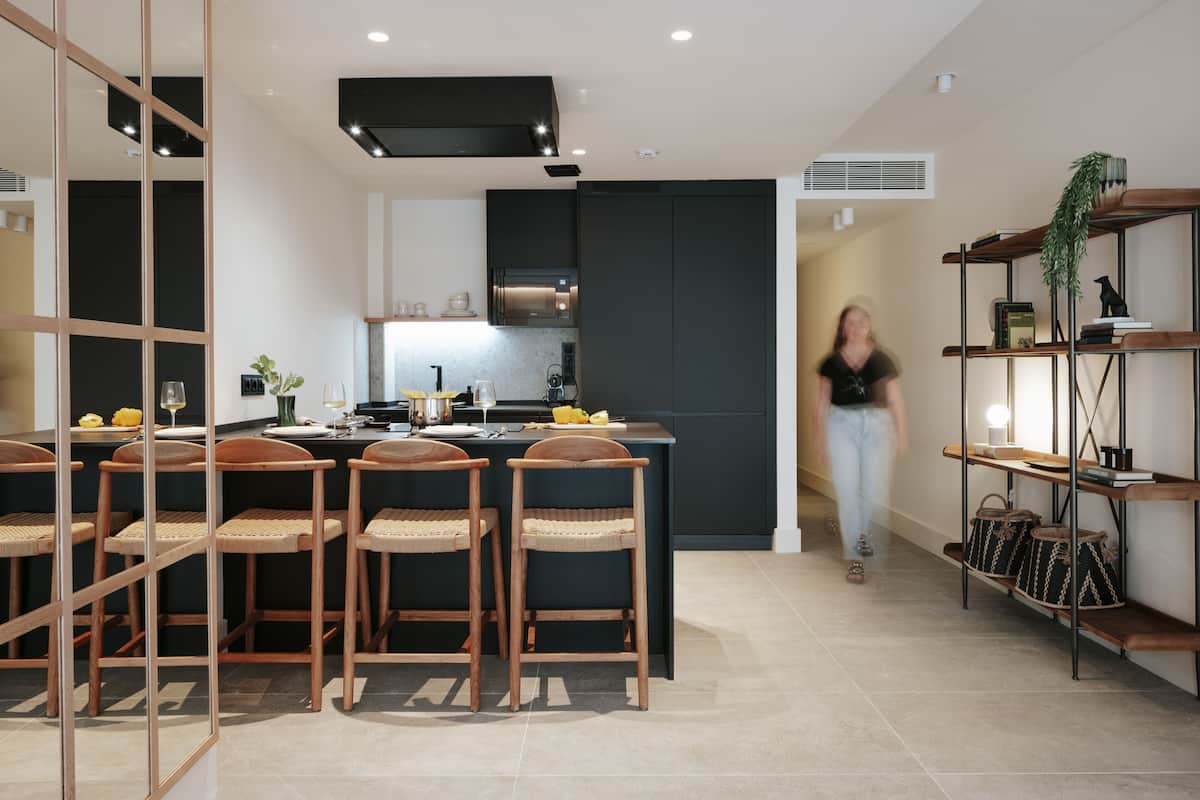 Une cuisine moderne avec des meubles de couleur sombre, un îlot central doté de tabourets de bar, et une personne que l'on aperçoit traversant l'espace.