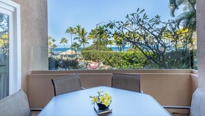 Un balcon avec une table et des chaises, un vase rempli de fleurs et une vue sur les palmiers et l’océan.