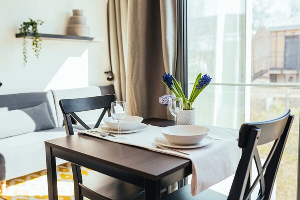 A dining area with a table set for two, a vase with flowers, and a view of the outside through a window.