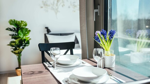 A dining table set with white plates, cutlery, and glasses, with a vase of blue flowers on the window sill.