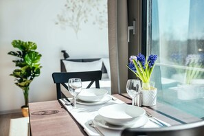 Mesa de jantar posta com pratos brancos, talheres e copos, com um vaso de flores azuis no parapeito da janela.