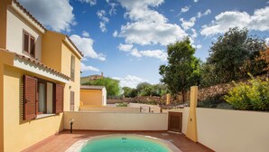 A swimming pool area with a tiled deck, a stone wall, and a building with a yellow facade and brown shutters. - Villa con Piscina - BUDONI (San Lorenzo)