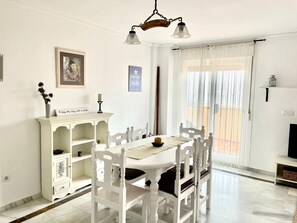 A dining area with a white table and chairs, a white cabinet with shelves, a chandelier, and a window with sheer curtains. - House (Bahía de Casares)