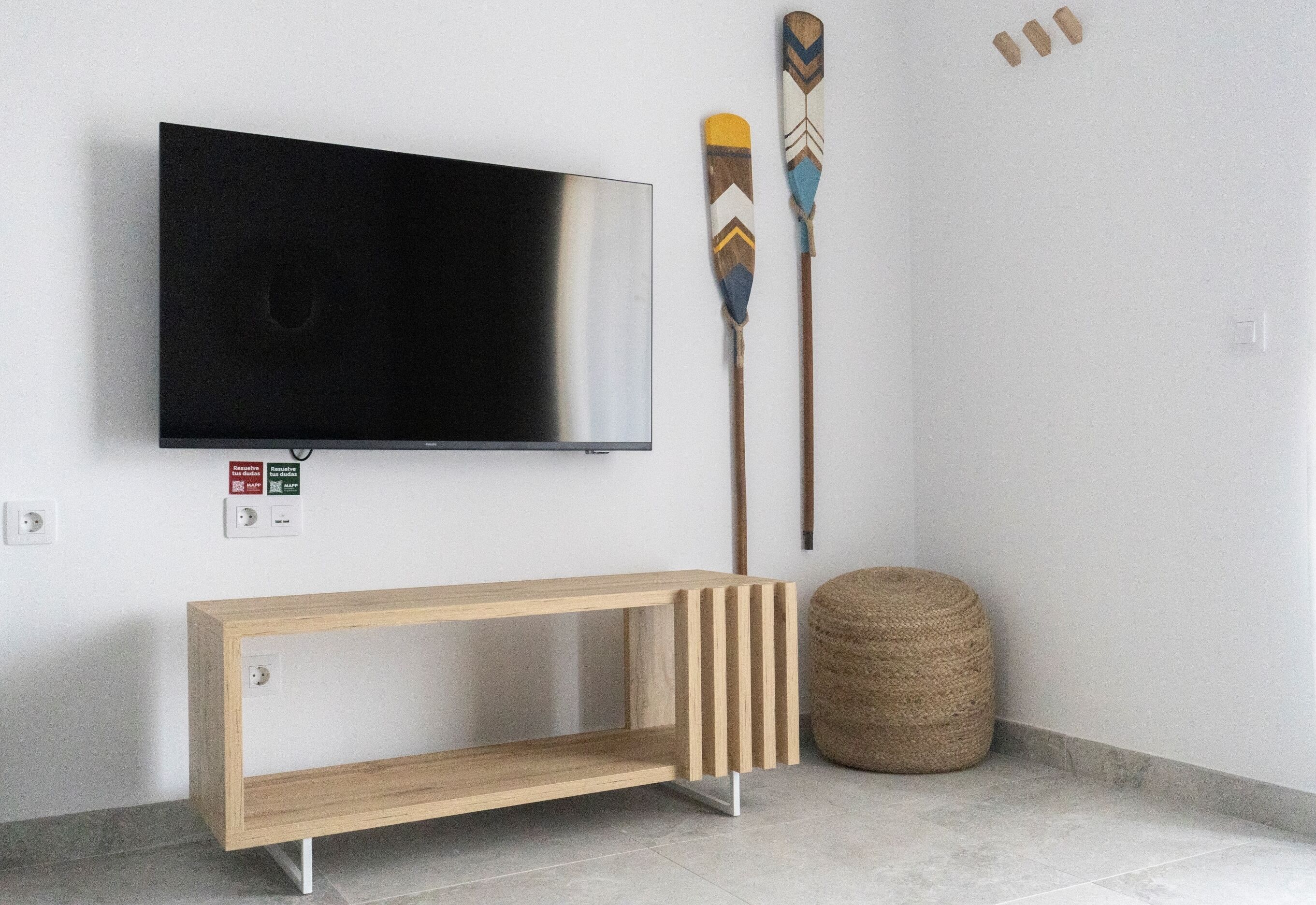 A modern living room with a flat-screen TV mounted on the wall, a wooden console table, two decorative paddles, and a woven ottoman.