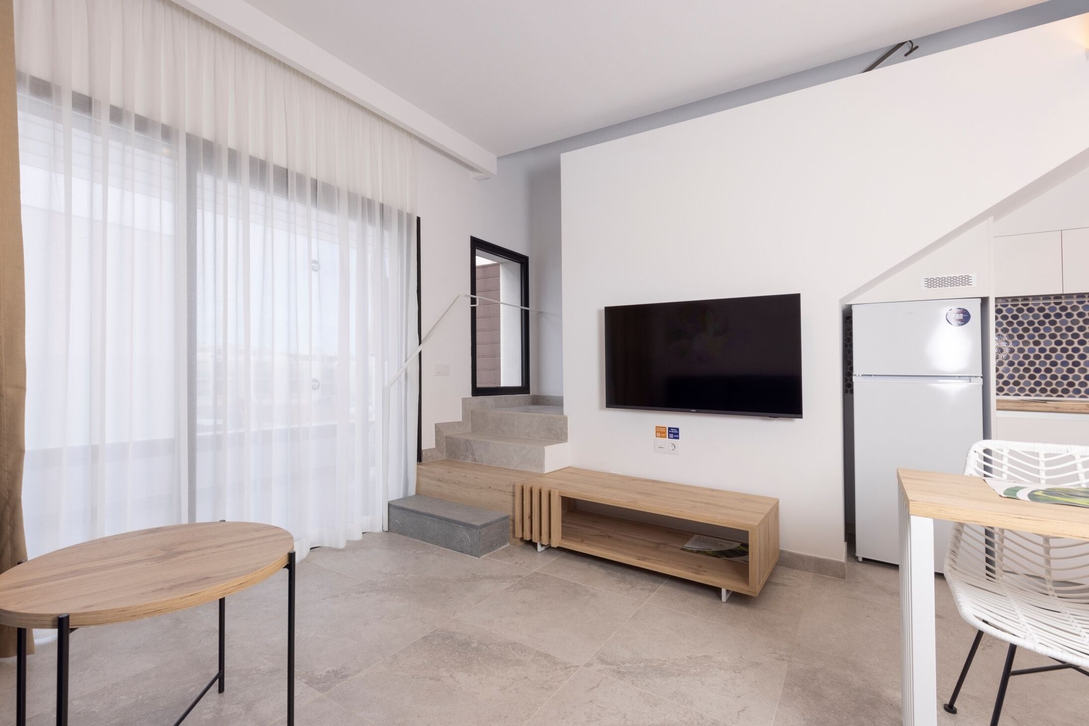 A modern living room with a flat-screen TV, a wooden bench, and a dining area with a white chair.