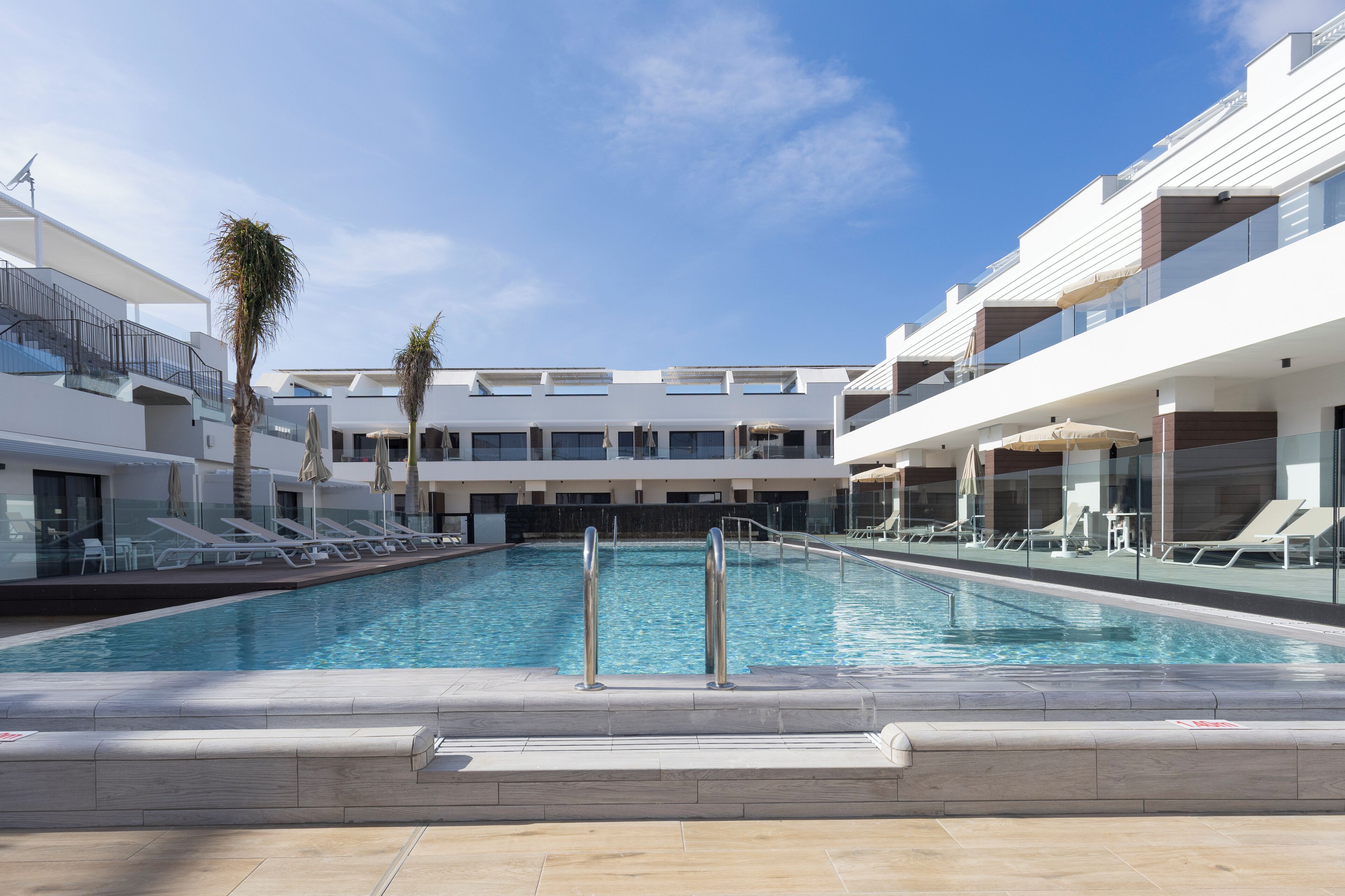 A modern hotel pool area with a clear blue pool, sun loungers, and a glass balustrade.