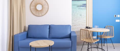 Una habitación con un sofá azul, una mesa redonda de madera, sillas de mimbre, un espejo montado en la pared y vista al océano a través de una ventana.