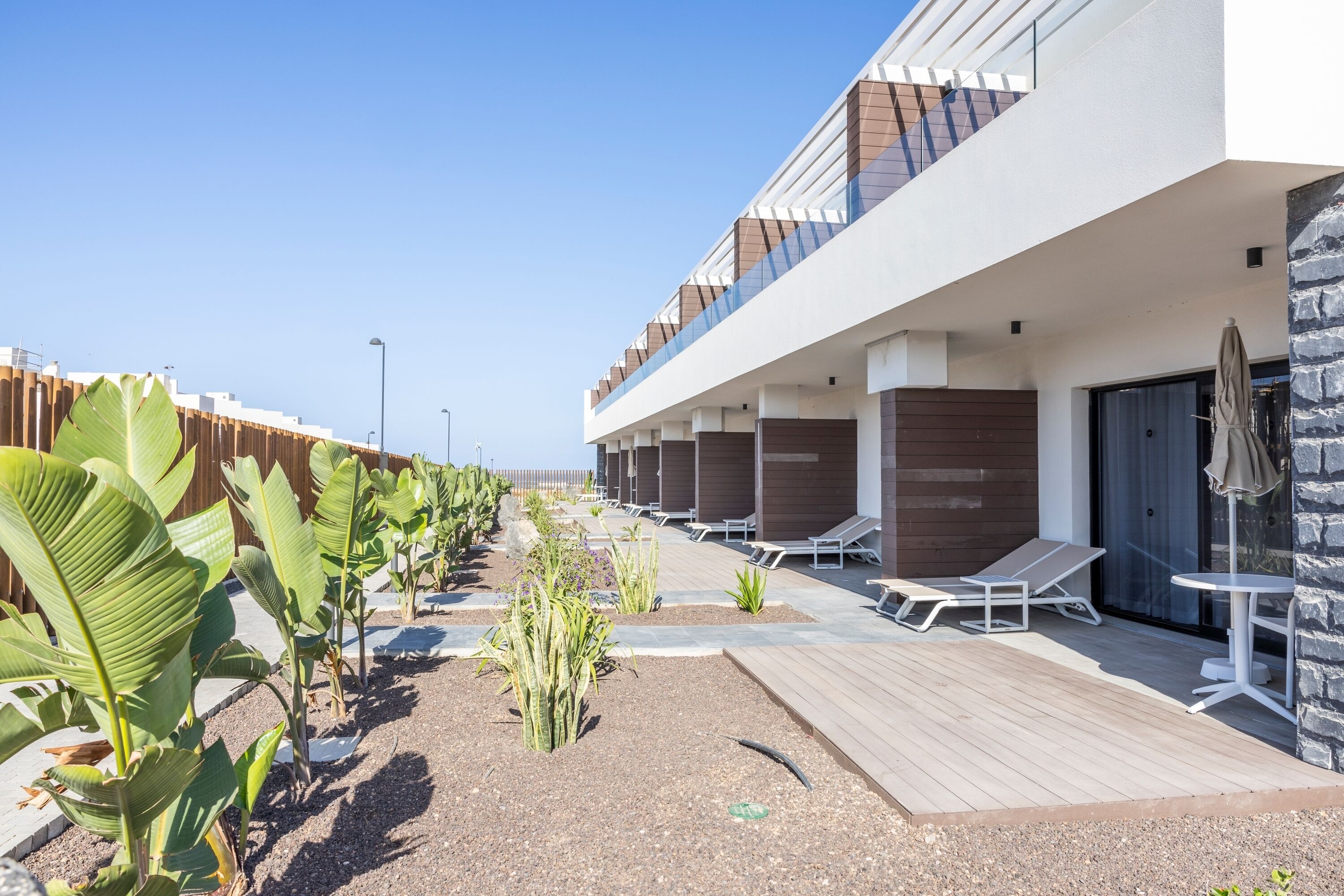 Ein modernes Hotel mit Holzterrasse, Liegestühlen und einer Reihe Zimmer mit großen Fenstern.