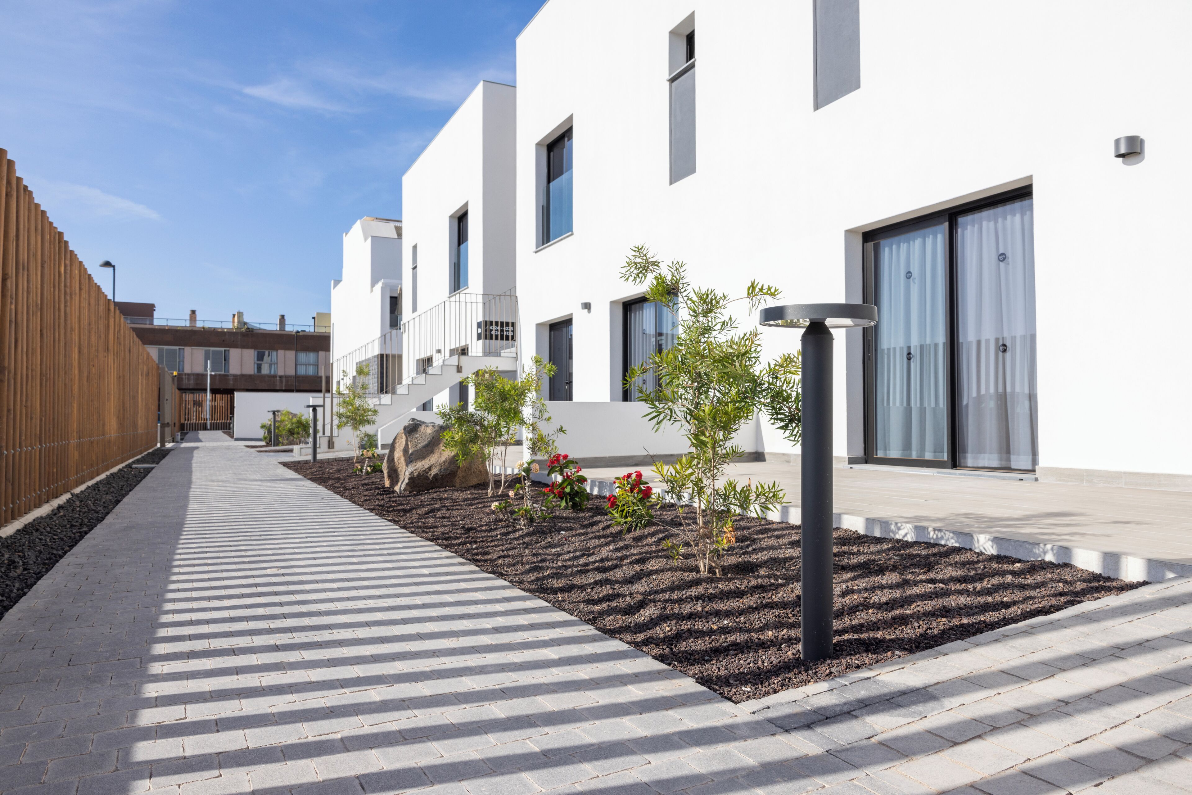 A modern building with a clean, white facade, a wooden fence on the left, and a landscaped area with small plants and a large rock.