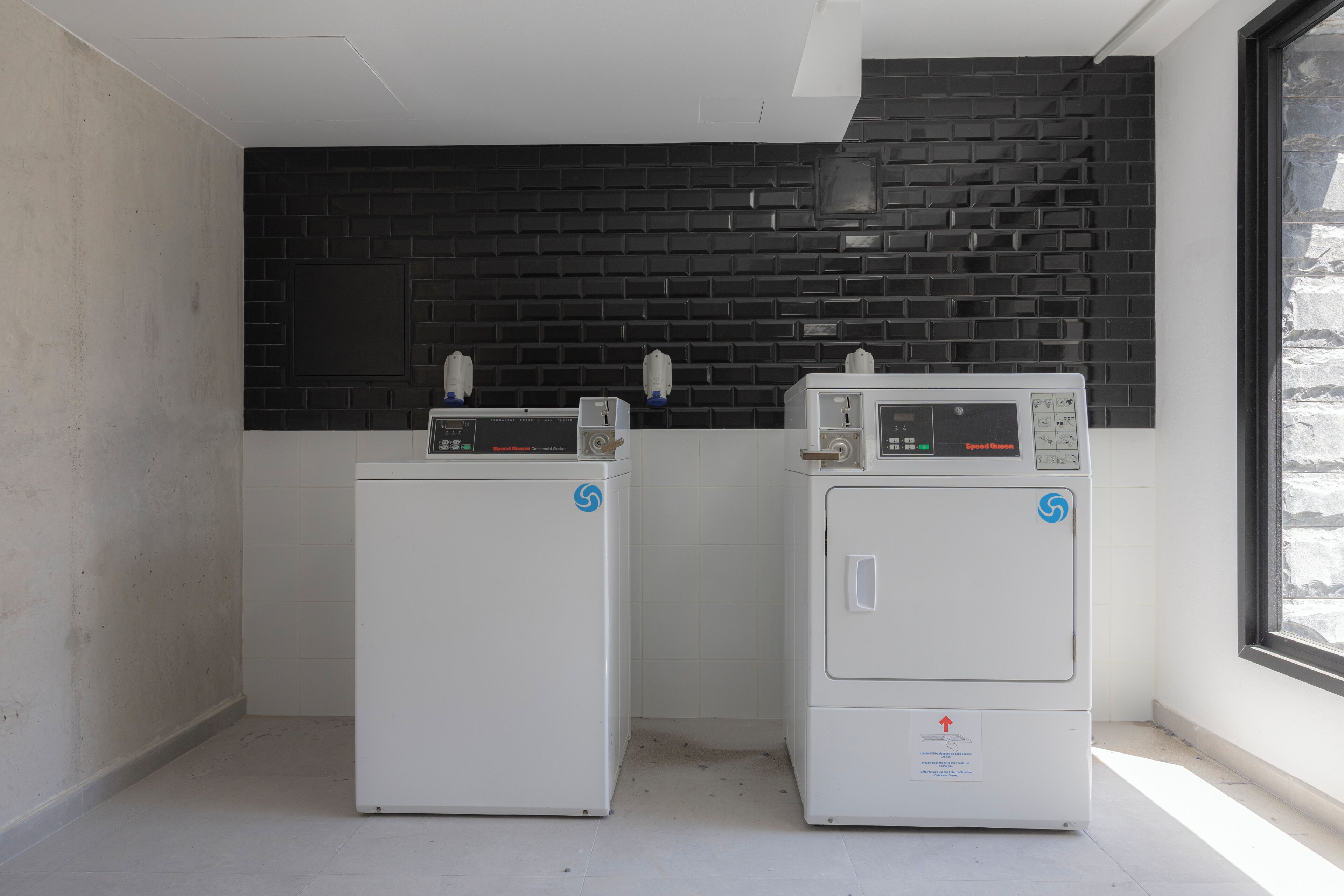 Two white washing machines in a corner with a black brick wall and a window.