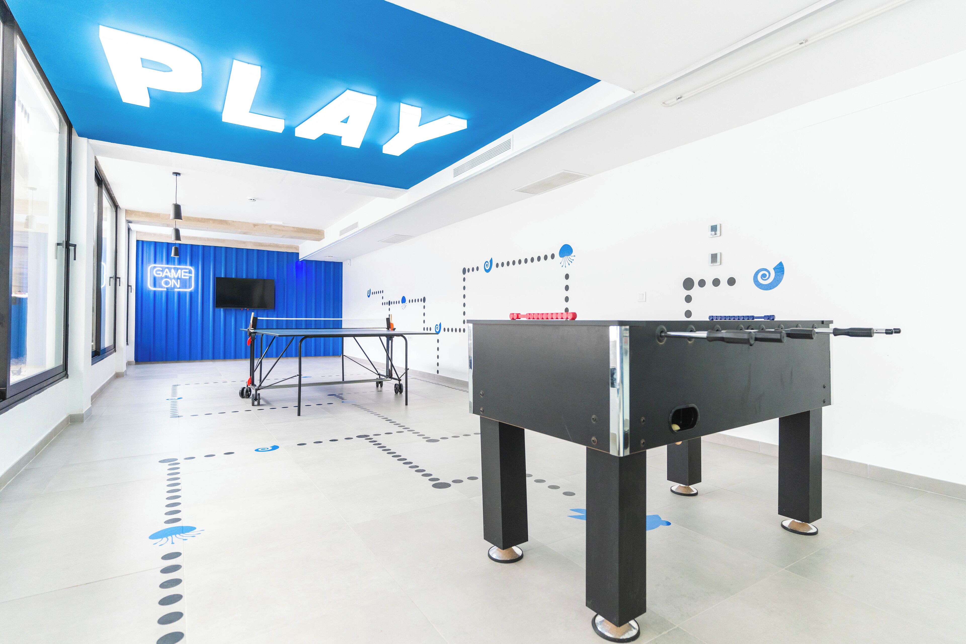 Un espacio interior moderno con una mesa de fútbol de mesa, una pared azul con un letrero que dice 'GAME ON' y un techo con letras iluminadas que dicen 'PLAY'.