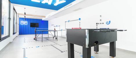 Un espacio interior moderno con una mesa de fútbol de mesa, una pared azul con un letrero que dice 'GAME ON' y un techo con letras iluminadas que dicen 'PLAY'.