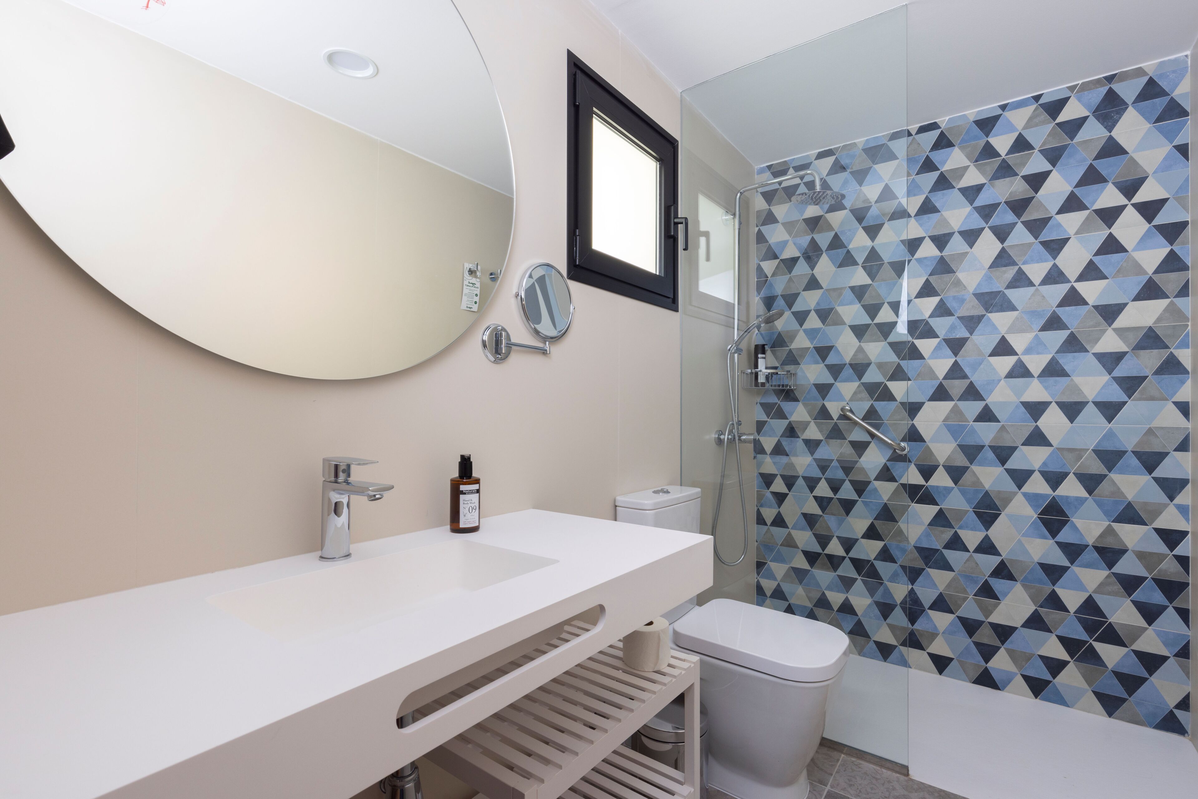 Un baño moderno con tocador blanco, un espejo grande, inodoro y ducha con azulejos de diseño geométrico.