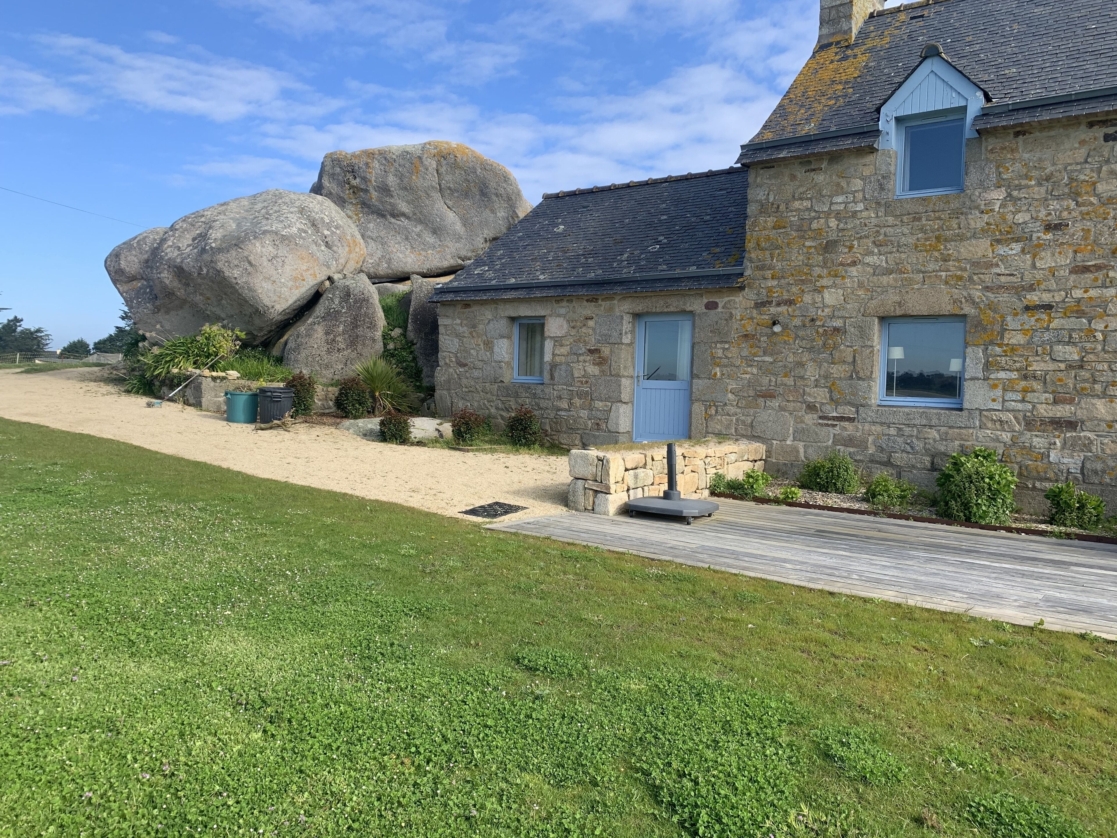 A stone house with a large rock formation nearby, a paved driveway, and a grassy lawn.