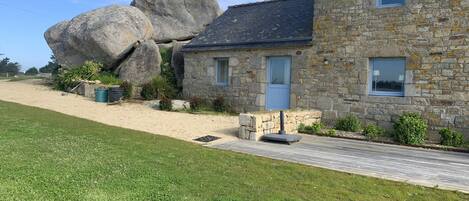 A stone house with a large rock formation nearby, a paved driveway, and a grassy lawn.