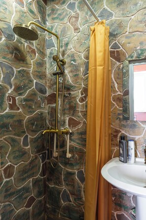 A bathroom with a stone wall, a shower with a gold showerhead, and a yellow shower curtain. - Etiuni The Forgotten Kingdom (Sevan)