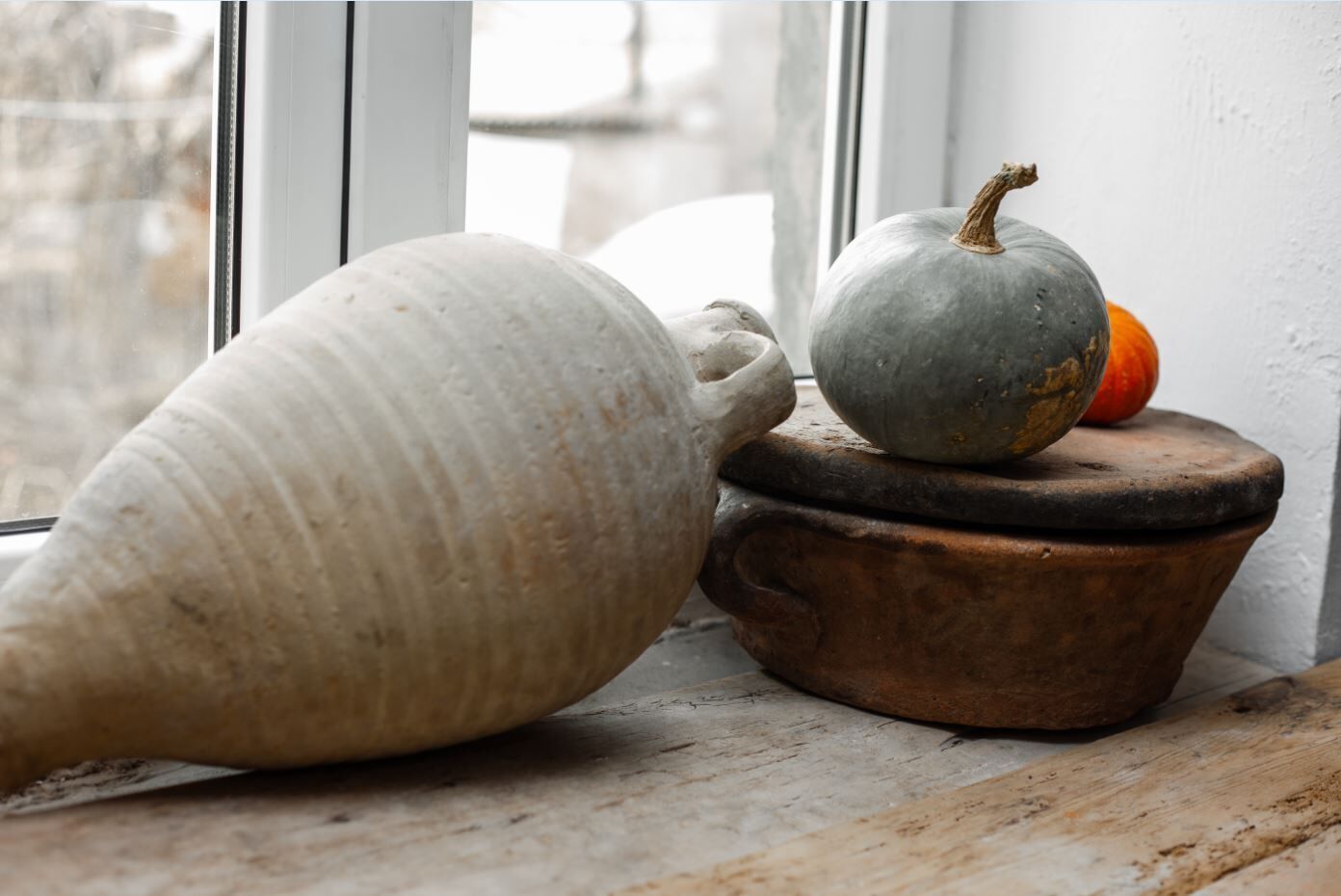 Un jarrón de cerámica grande y una pequeña calabaza sobre una mesa de madera rústica cerca de una ventana.