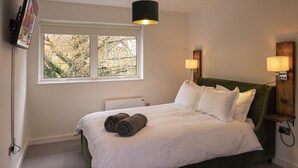 Una cama bien tendida con sábanas blancas y dos toallas enrolladas, una ventana con vistas a árboles, y dos lámparas fijadas a la pared.