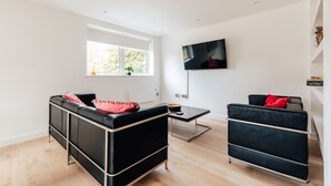 A modern living room with a black leather sofa, a black armchair, and a glass coffee table. There is a flat-screen TV mounted on the wall and a window providing natural light. - Luxury 2 Bed/2 Shower Room Apartment in Bath City (Bath)